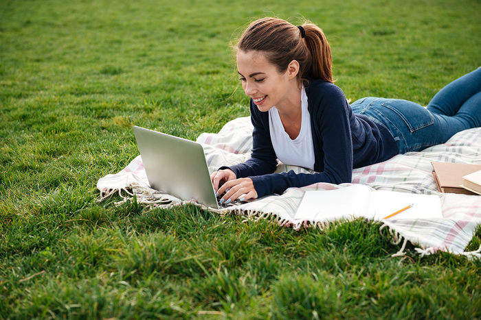 Happy teenage girl laying on blanket outside on the grass, typing on a laptop.